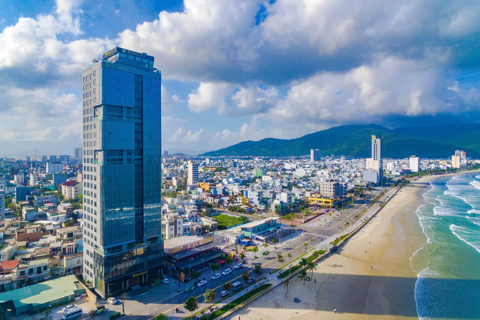 High-rise building of Peninsula Hotel Danang on the coast next to a sandy beach.