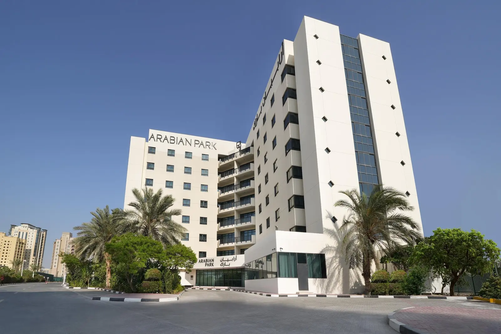 White building of Arabian Park Dubai hotel with palm trees and a driveway on a sunny day.