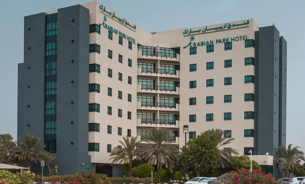 Facade of the multi-story Arabian Park hotel building in Dubai behind green trees.